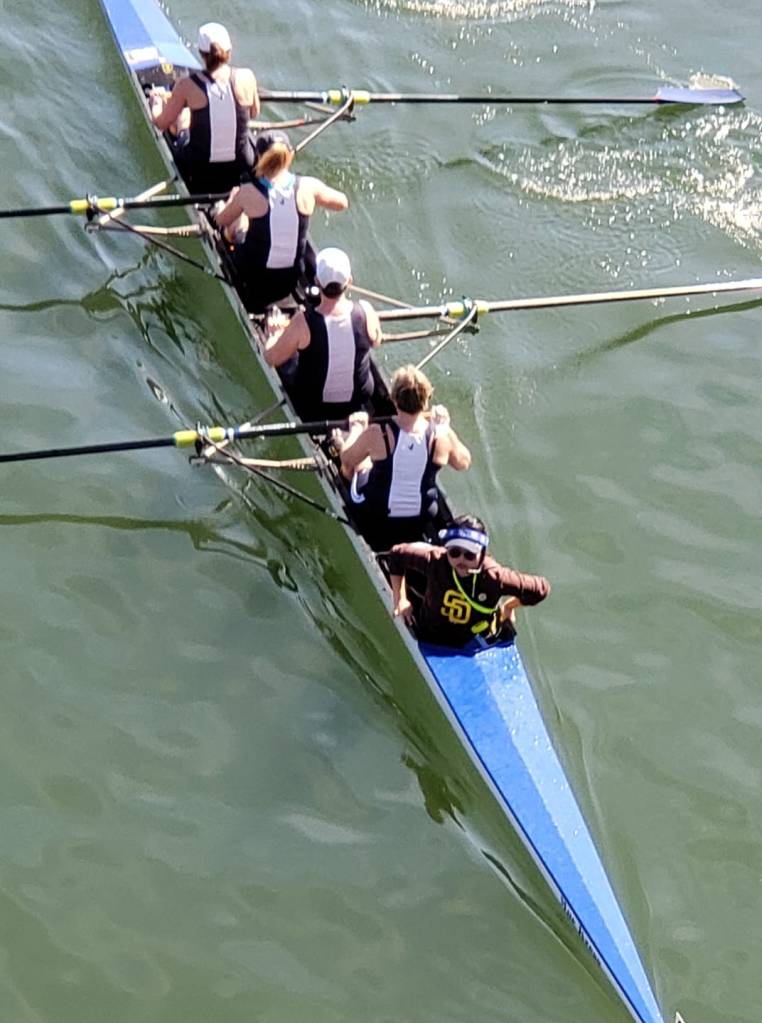 Four rowers facing backward and one coxswains facing forward in a boat on the water.