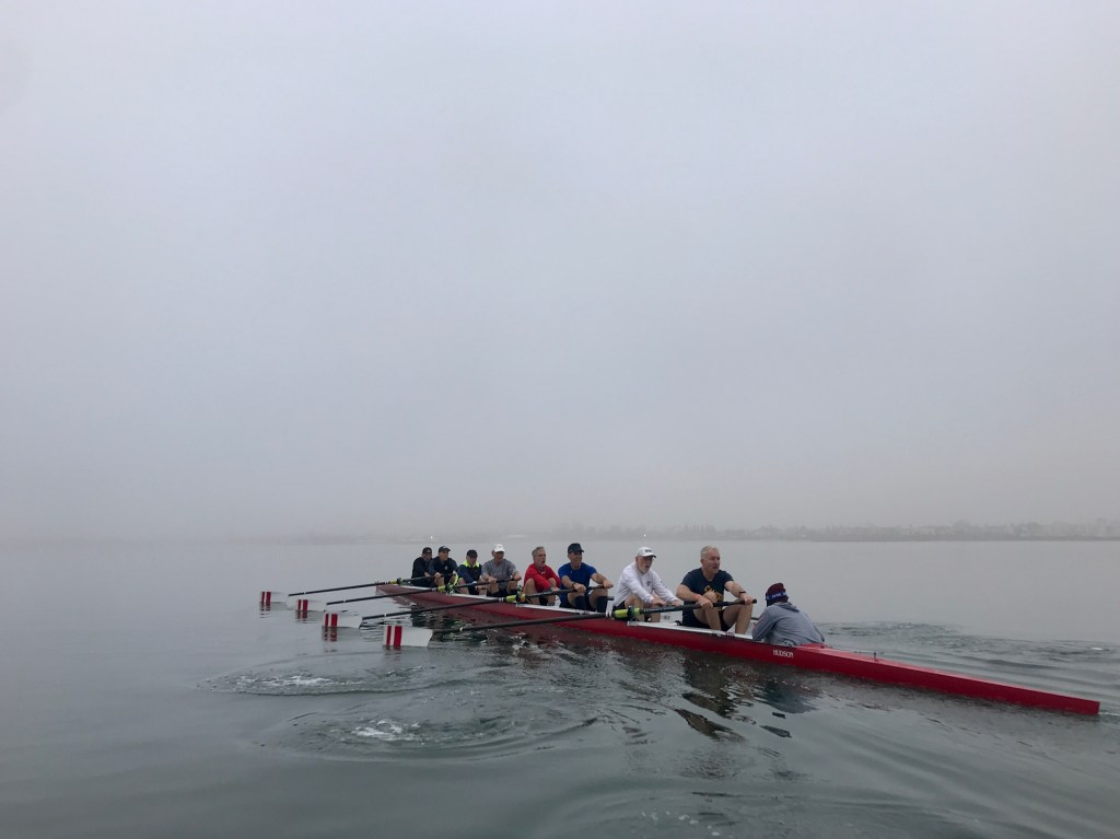 Eight rowers and a coxswain in a red rowing shell with white blades with two red stripes being placed in the water. Foggy morning on Mission Bay.
